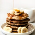 Banana blender pancake stack on a white plate with bananas and a drizzle of maple syrup on top.