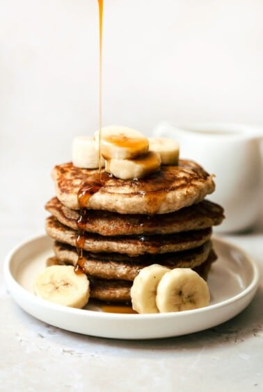 Banana blender pancake stack on a white plate with bananas and a drizzle of maple syrup on top.