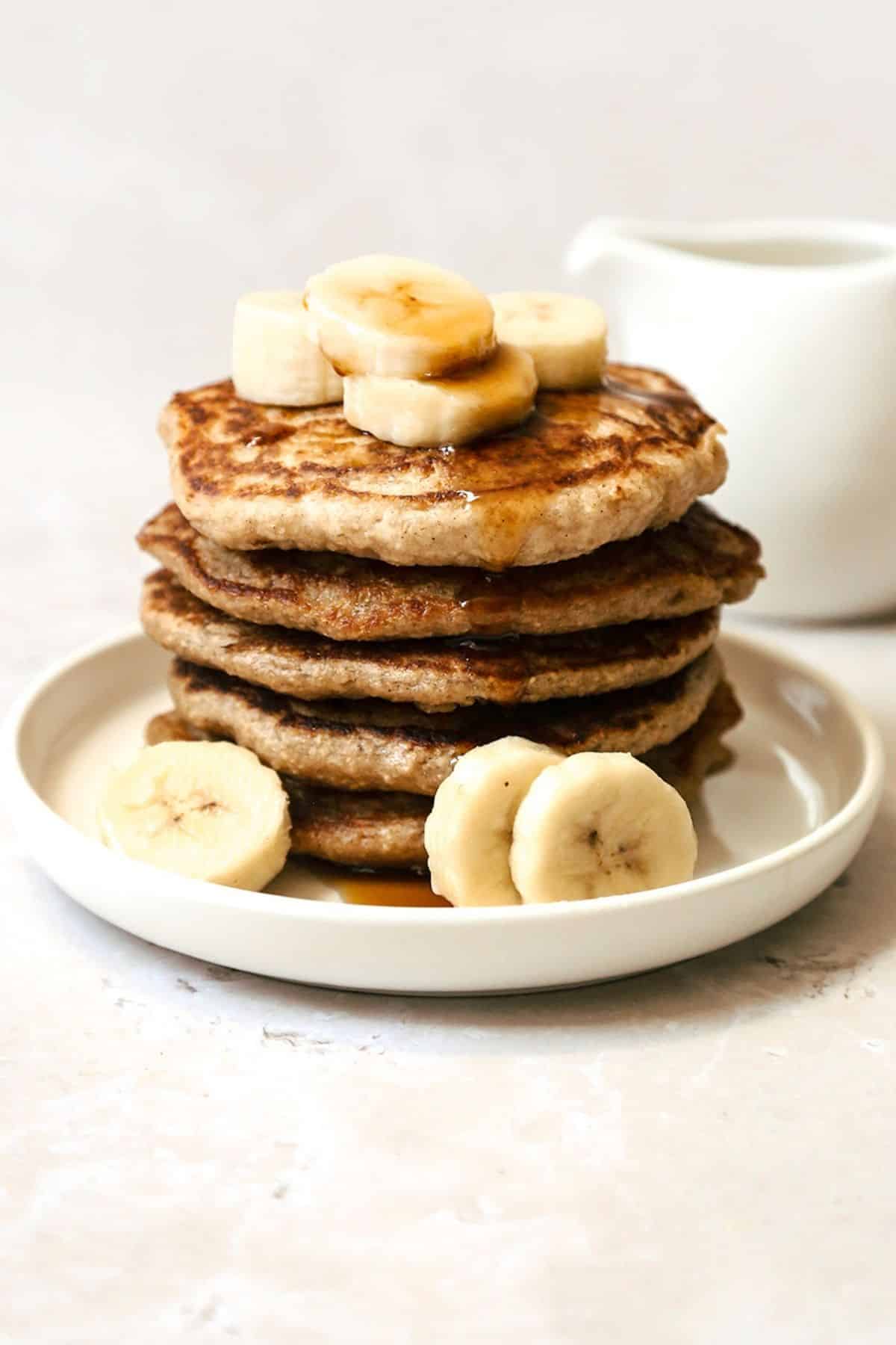 Close-up of banana oatmeal pancake stack on a white plate with bananas and a drizzle of maple syrup on top.