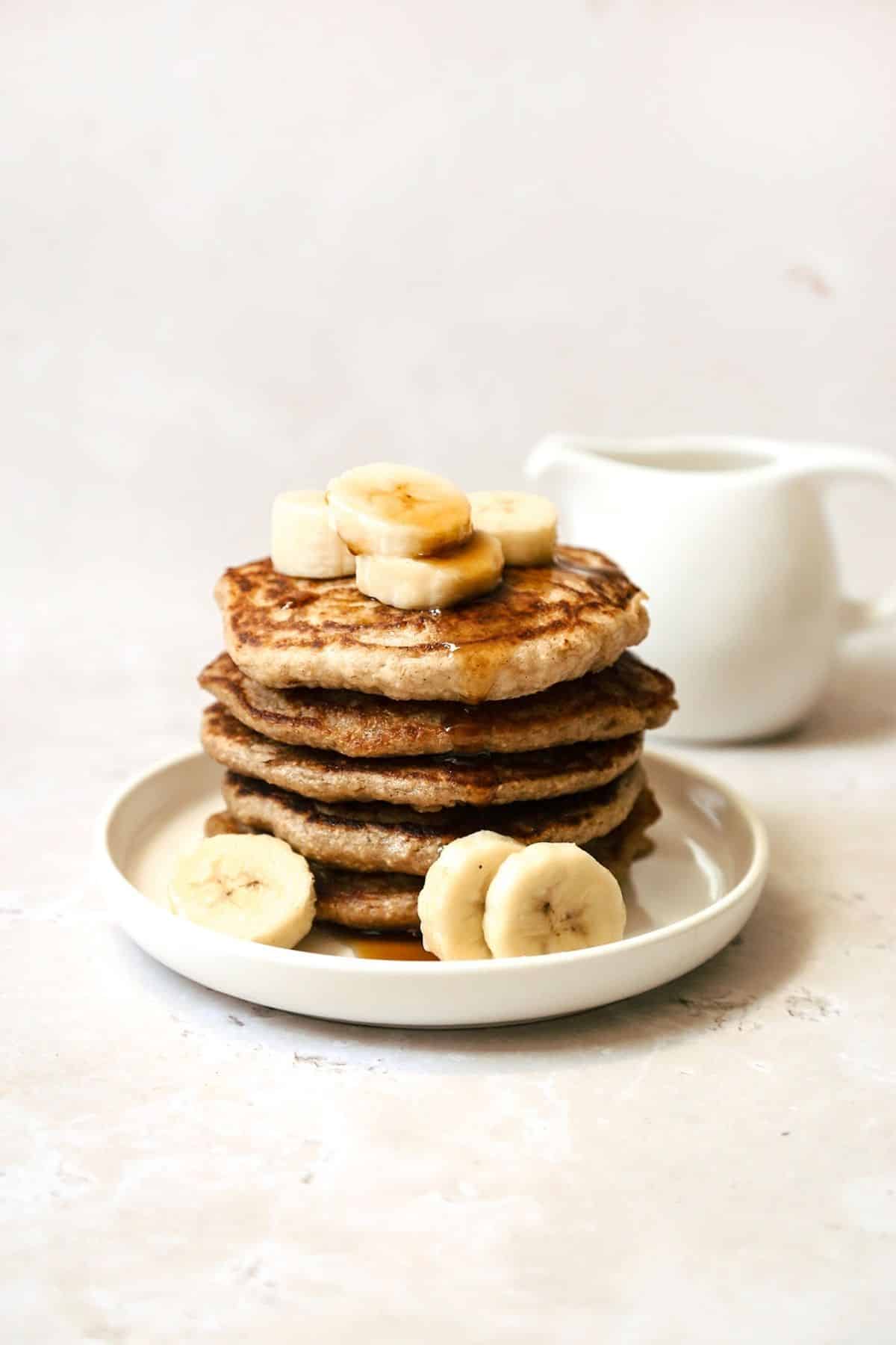 Banana oatmeal pancake stack on a white plate with bananas and a drizzle of maple syrup on top.