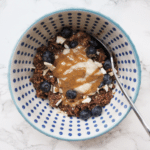 Chocolate oats in a blue patterned bowl with a marble backdrop, with yogurt, blueberries. coconut and peanut butter drizzled on top. Stainless steel spoon scooping the oats.