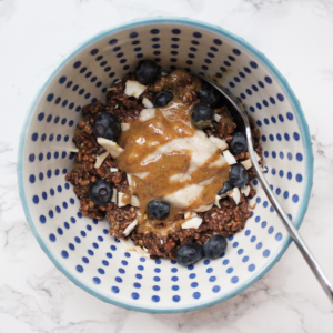 Chocolate oats in a blue patterned bowl with a marble backdrop, with yogurt, blueberries. coconut and peanut butter drizzled on top. Stainless steel spoon scooping the oats.