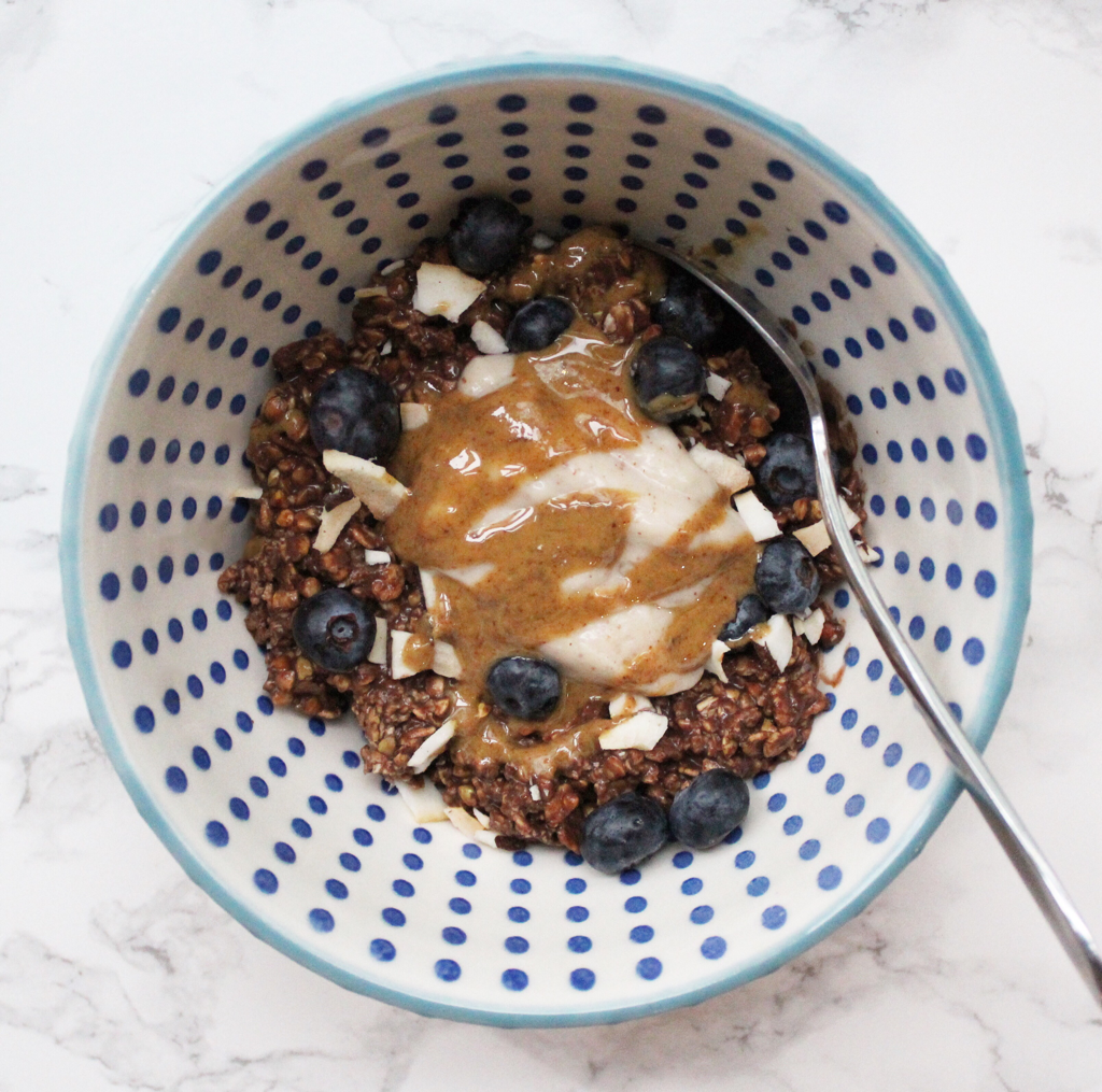 Chocolate oats in a blue patterned bowl with a marble backdrop, with yogurt, blueberries. coconut and peanut butter drizzled on top. Stainless steel spoon scooping the oats.