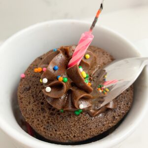 Chocolate mug cake with chocolate icing, sprinkles, a candle and a spoon taking a scoop of it.