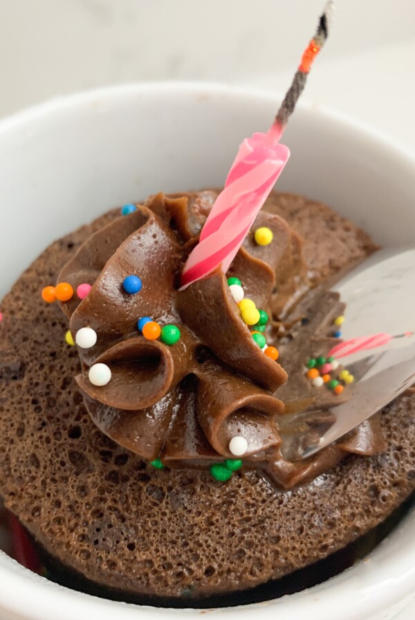 Chocolate mug cake with chocolate icing, sprinkles, a candle and a spoon taking a scoop of it.