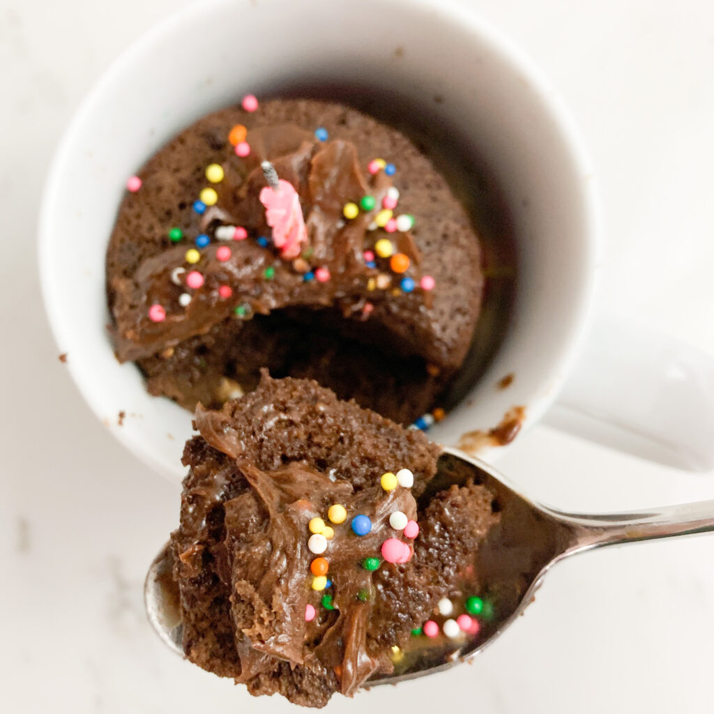 Chocolate mug cake with chocolate icing, sprinkles, a candle and a spoonful taken out of it.