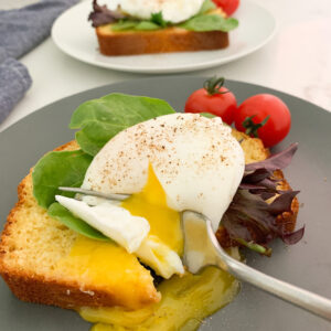 Cornbread with a poached egg and lettuce on top with a fork breaking the yolk on a blue plate. Another piece of cornbread with an egg on it is in the background.