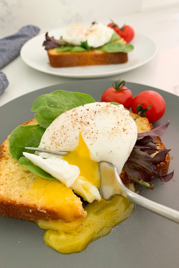 Cornbread with a poached egg and lettuce on top with a fork breaking the yolk on a blue plate. Another piece of cornbread with an egg on it is in the background.