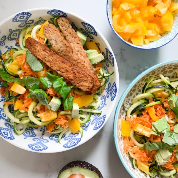Two tempeh bowls with zucchini, shredded carrots, sliced yellow pepper, avocado and basil. Props include small bowl with yellow bell pepper to the top right, and half of an avocado to the bottom.
