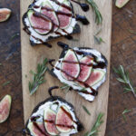 Three pieces of charcoal bread with cream cheese, sliced figs and balsamic reduction on a wooden plank. Rosemary sprinkled around them as garnish.