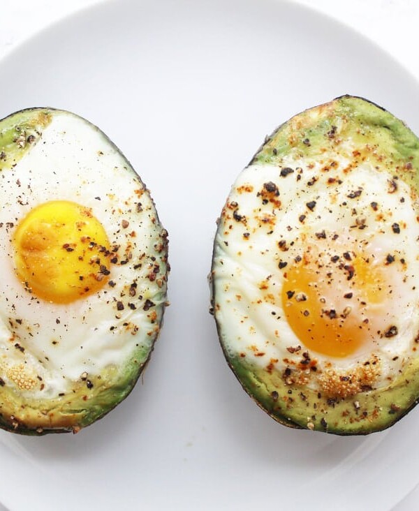 Two baked avocado halves with eggs and pepper on a white plate with a fork to the left and a knife to the right.