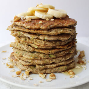 Stack of Banana Zucchini Pancakes with oats, walnuts, sliced bananas and syrup on top.