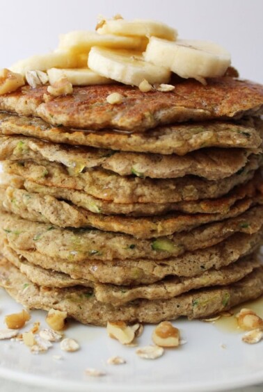Stack of Banana Zucchini Pancakes with oats, walnuts, sliced bananas and syrup on top.