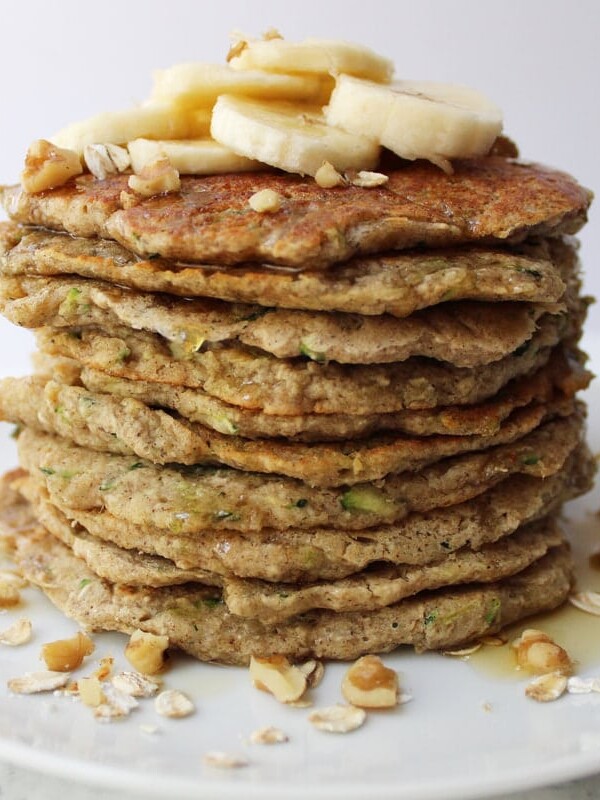 Stack of Banana Zucchini Pancakes with oats, walnuts, sliced bananas and syrup on top.