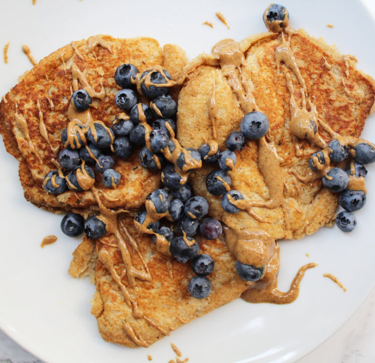Three banana pancakes on a white plate, with blueberries and drizzled with peanut butter.