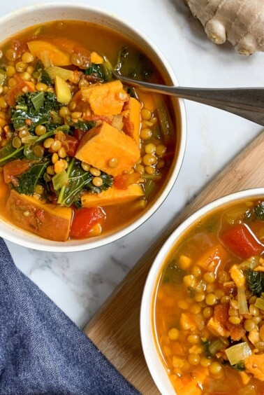 Two bowls of sweet potato, lentil and kale stew with a spoon. Blue napkin to left and fresh ginger in top right corner.
