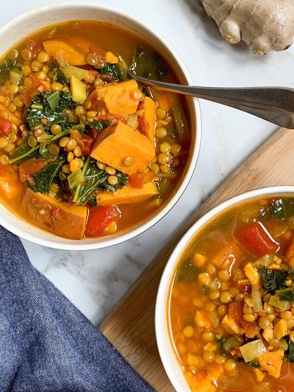 Two bowls of sweet potato, lentil and kale stew with a spoon. Blue napkin to left and fresh ginger in top right corner.