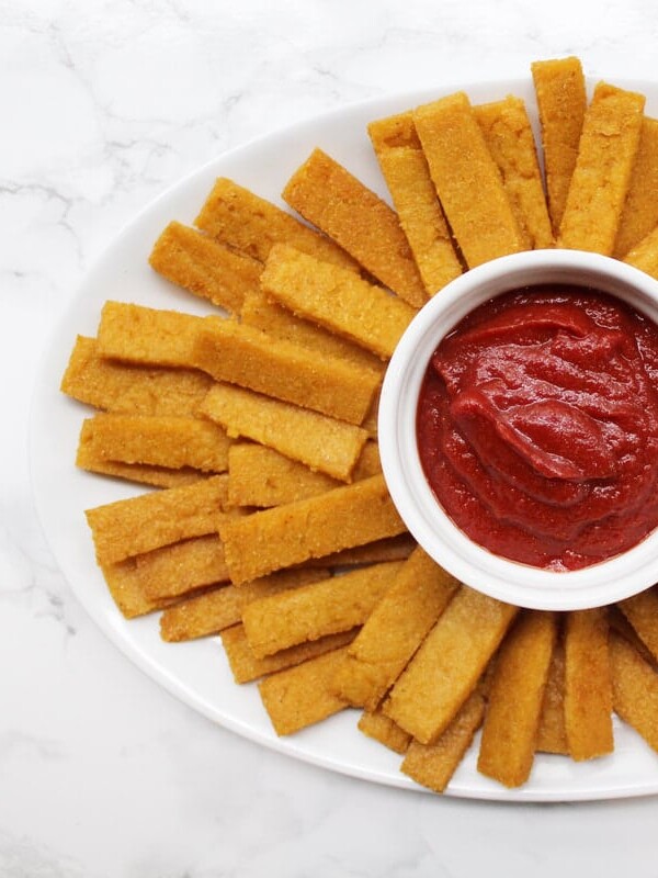 Baked polenta fries on a white plate with ketchup in the middle.