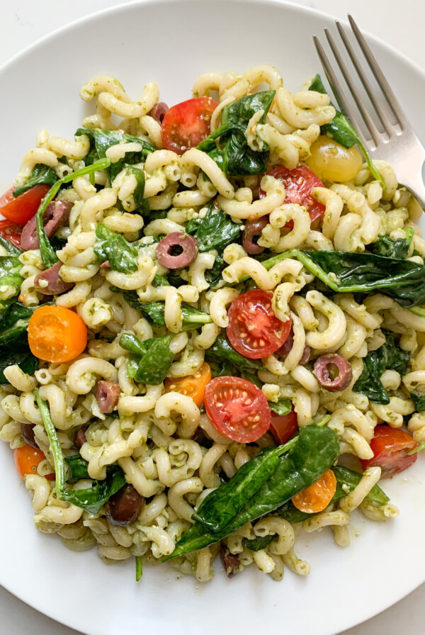 White plate filled with pasta salad - including brown rice pasta, pesto, spinach, tomatoes and black olives - with a fork on the top right.