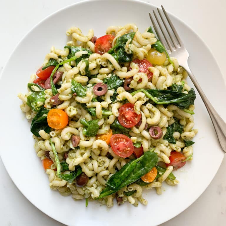 White plate filled with pasta salad - including brown rice pasta, pesto, spinach, tomatoes and black olives - with a fork on the top right.