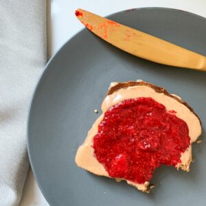 Piece of toast with peanut butter and strawberry jam on a blue plate with a gold-coloured butter knife. Light grey napkin to the left of the plate.