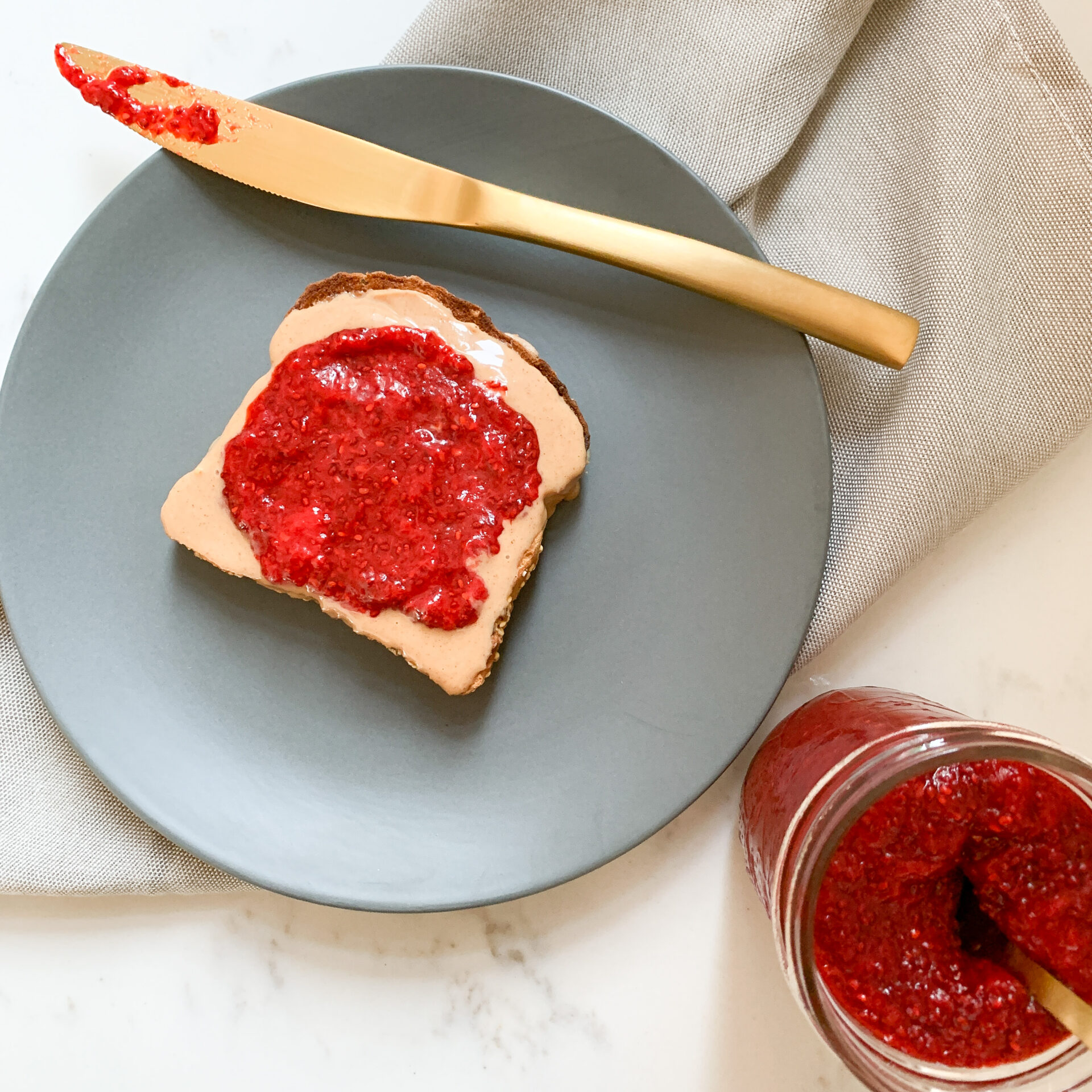 Piece of toast with peanut butter and strawberry jam on a blue plate with a gold-coloured butter knife. Light grey napkin under the plate and a jar of jam to the bottom right of the plate.