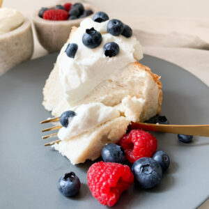 Angel food cake with whipped cream, blueberries and raspberries on a blue plate with a brass fork cutting into it. Berries and whipped cream bowls behind the cake.