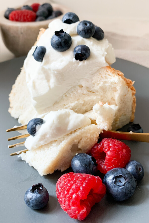 Angel food cake with whipped cream, blueberries and raspberries on a blue plate with a brass fork cutting into it. Berries and whipped cream bowls behind the cake.