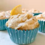 Lemon muffin with glaze in blue muffin cup. Lemon slice on top and other muffins behind.