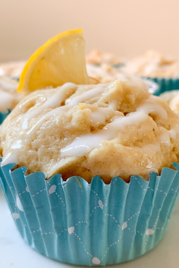 Lemon muffin with glaze in blue muffin cup. Lemon slice on top and other muffins behind.