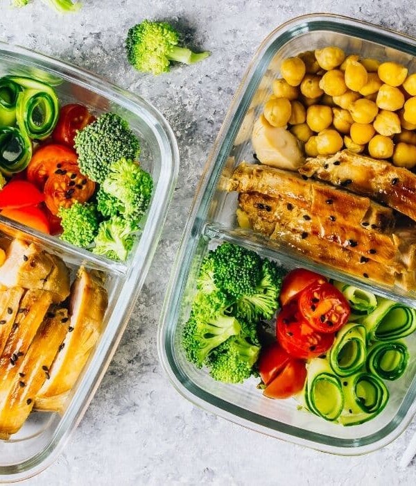 Two glass containers with chickpeas, chicken, tomatoes, zucchini and broccoli on grey tabletop. Fork and knife off to right.
