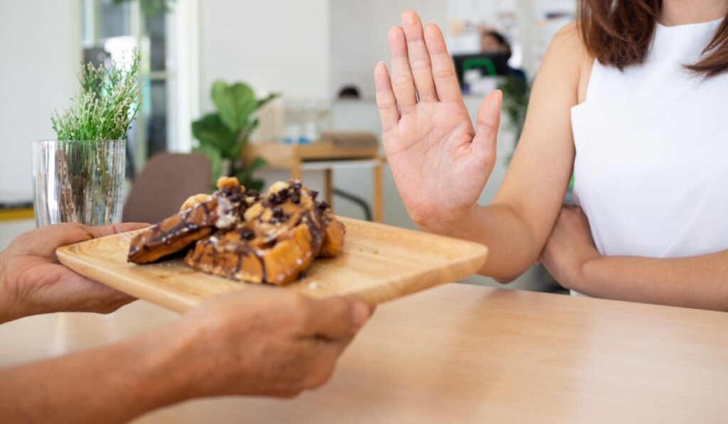 Woman in white shirt putting hand out to push away plate of waffles being handed to her.