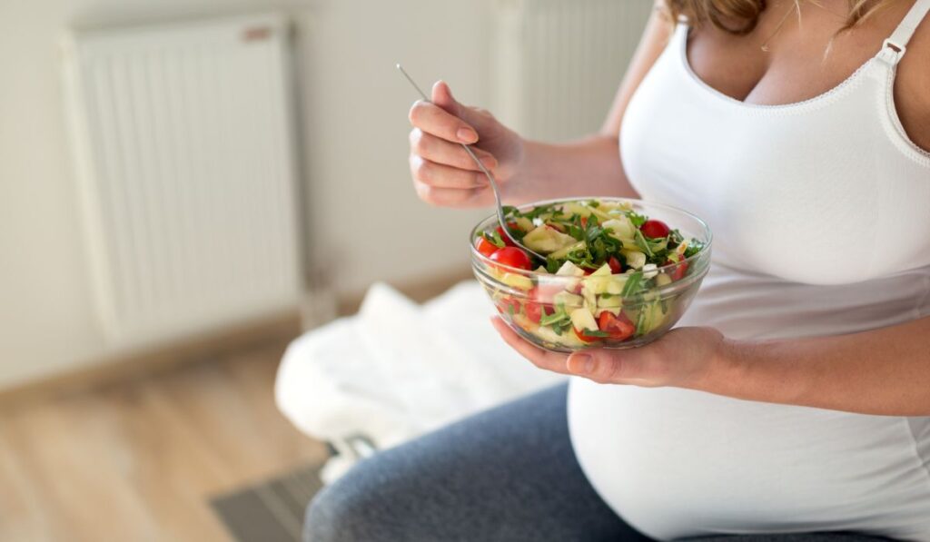 Pregnant woman sitting eating a bowl of salad.