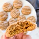 Almond flour pumpkin cookies on a plate and one cookie being held to camera with a bite taken out of it with a blue napkin in the right corner.