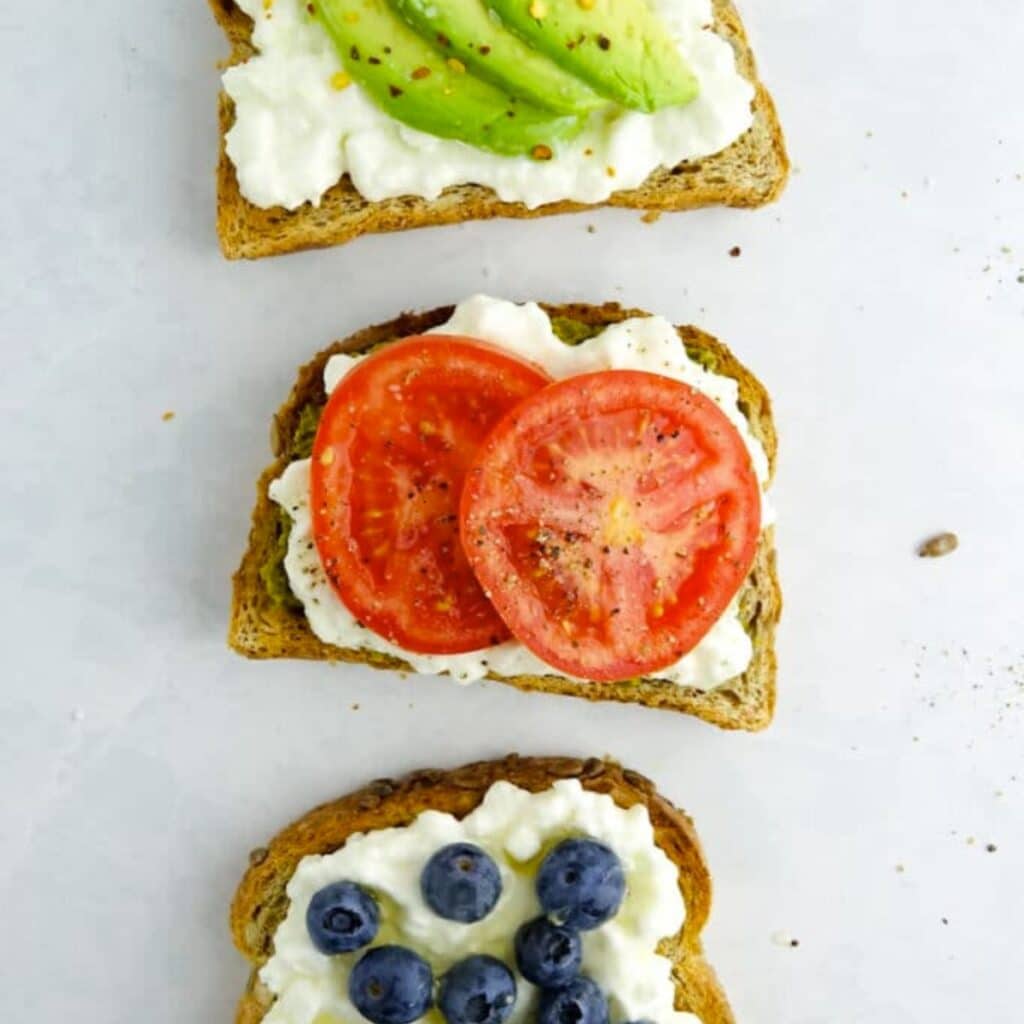 Overhead image of three pieces of cottage cheese toast. One topped with avocado, one with tomato and one with blueberries.