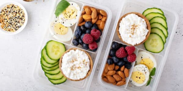 Two snack boxes with fruit, nuts, eggs, cucumber, and cottage cheese.