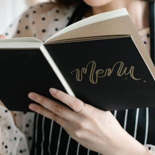Person in white polka dot shirt and apron writing in black book that says "menu".