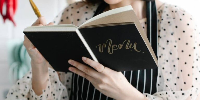 Person in white polka dot shirt and apron writing in black book that says "menu".