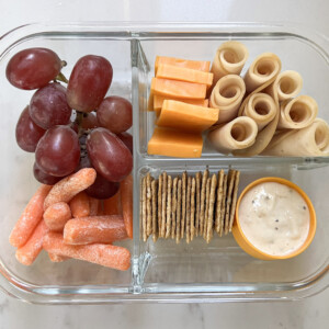 Glass lunch box with grapes, carrots, dip, crackers, cheese, and chicken deli meat.