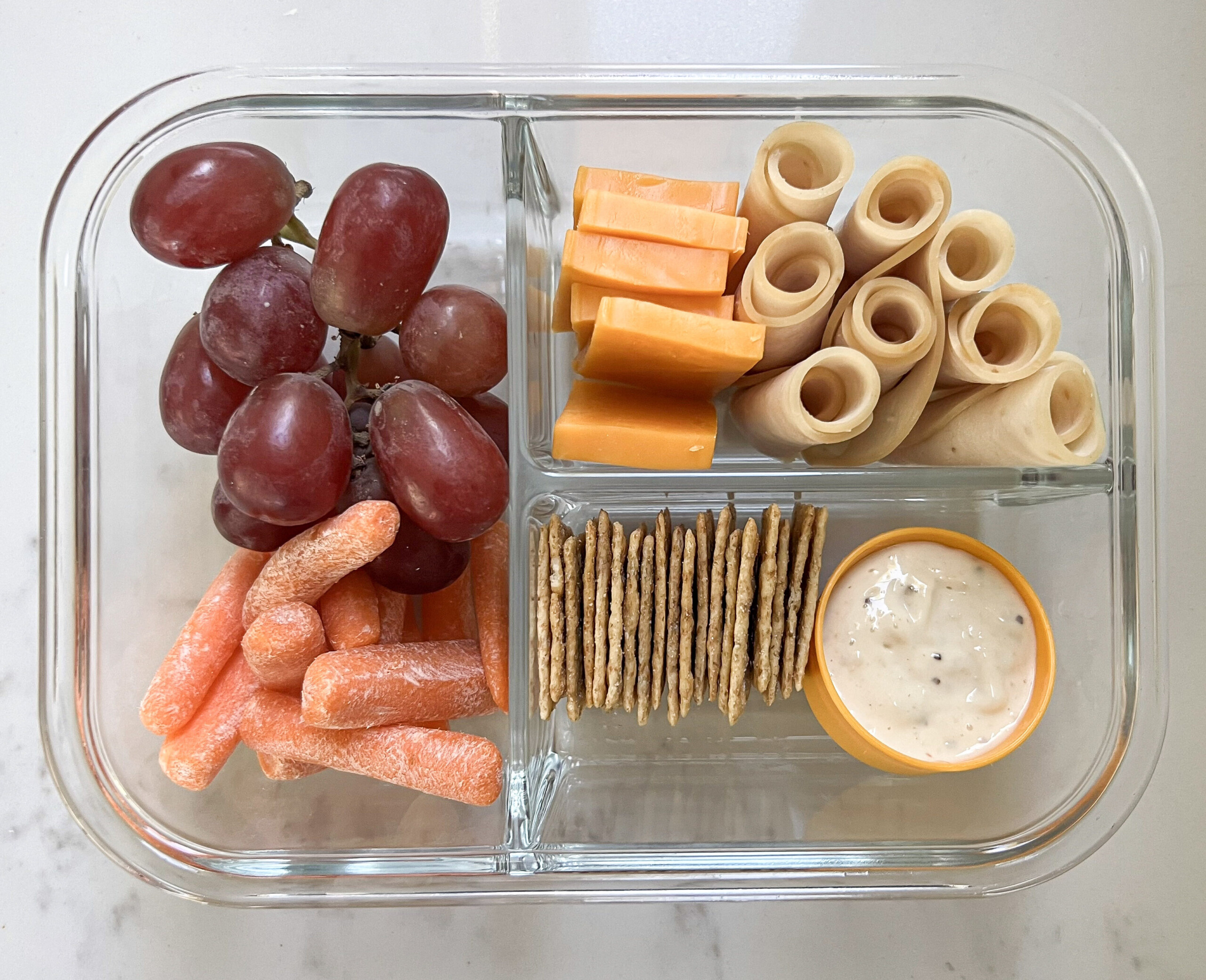 Glass lunch box with grapes, carrots, dip, crackers, cheese, and chicken deli meat.