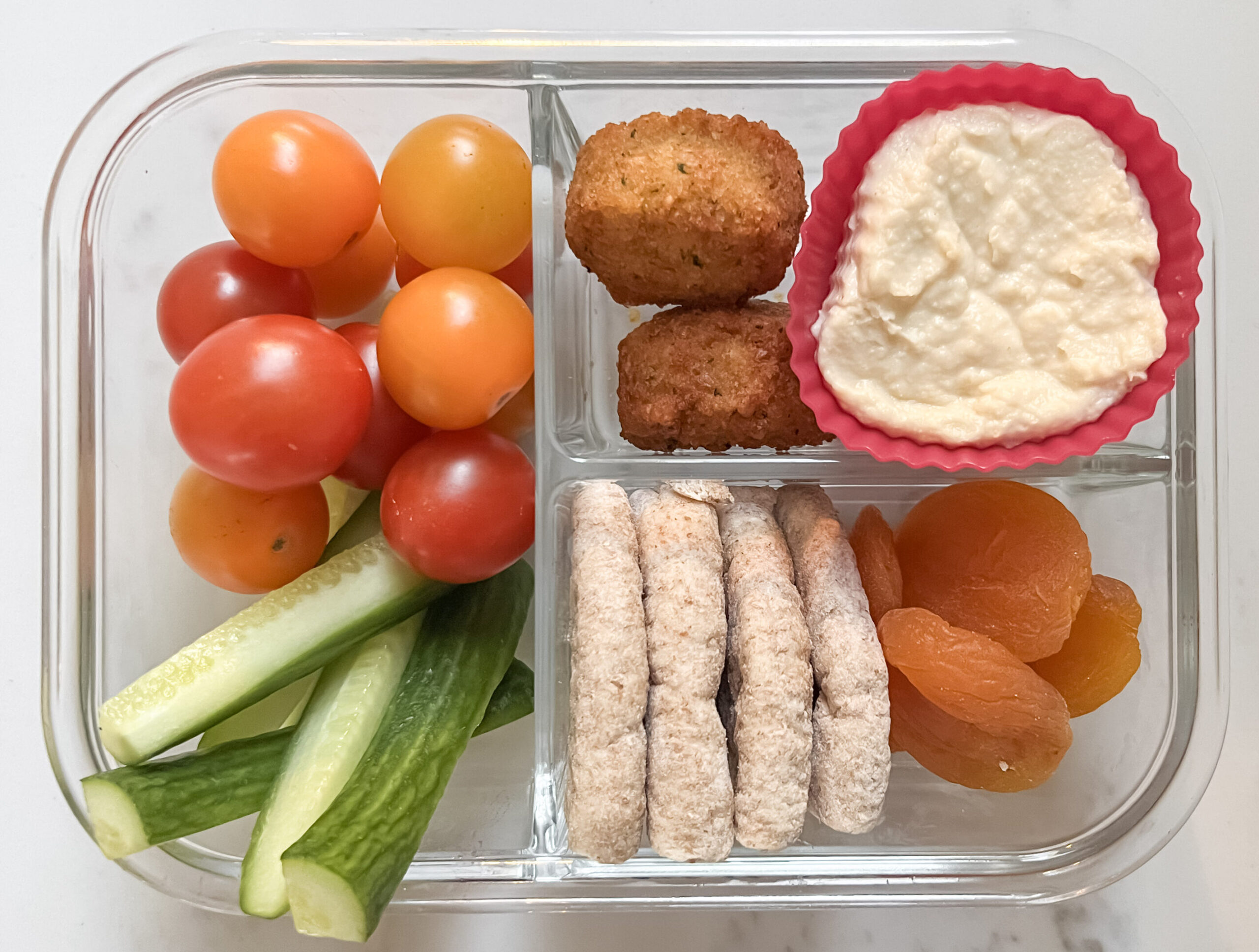 Glass lunch box with tomatoes, cucumber, pita, falafel, apricots, and hummus.
