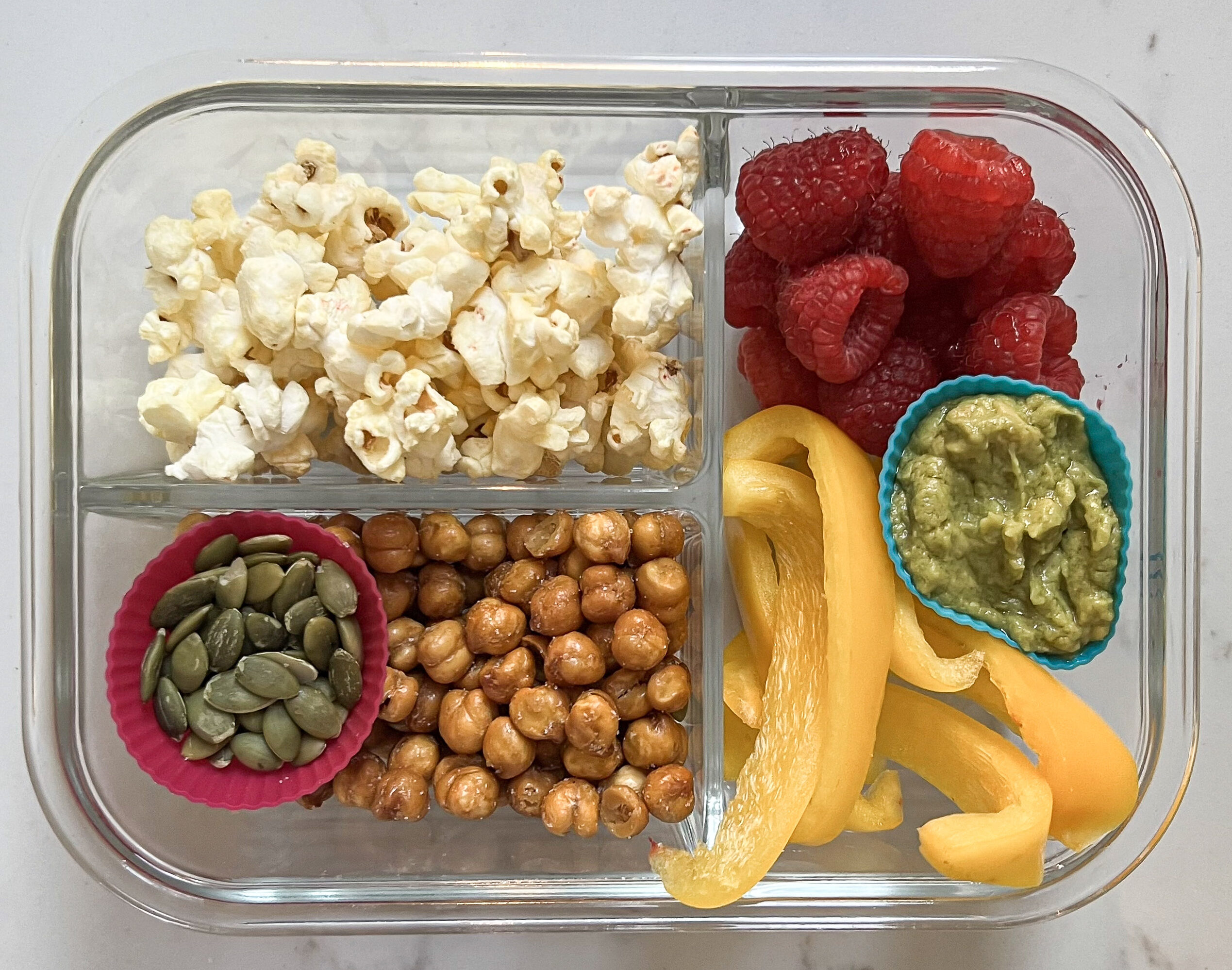 Glass lunch box with popcorn, raspberries, peppers, guacamole, chickpeas, and pumpkin seeds.