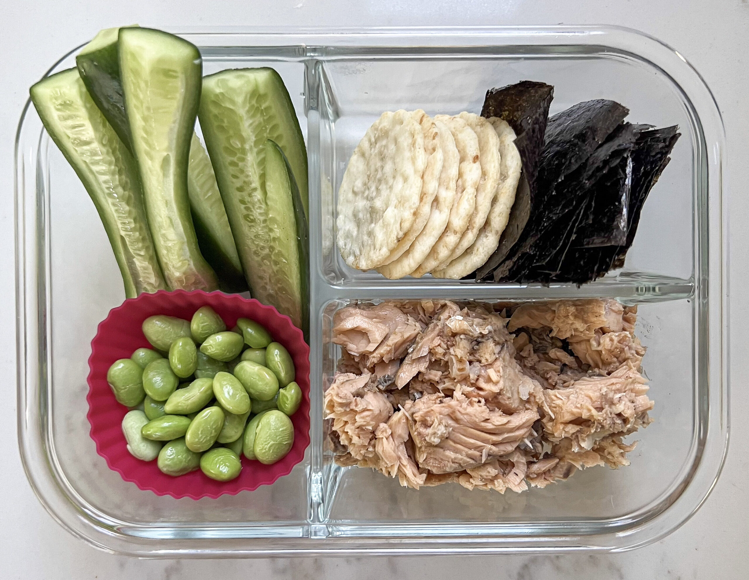 Glass lunch box with cucumber, edamame, rice crackers, nori, and canned salmon.