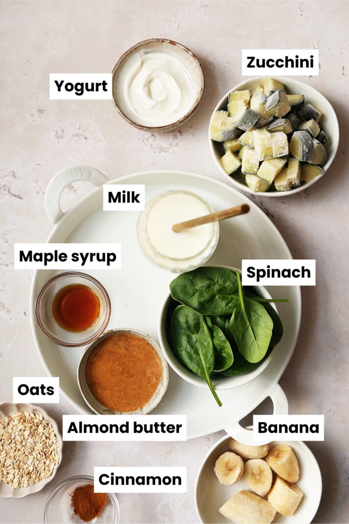 Overhead shot of zucchini smoothie ingredients, including yogurt, frozen zucchini, milk, spinach, maple syrup, almond butter, oats, cinnamon and banana.