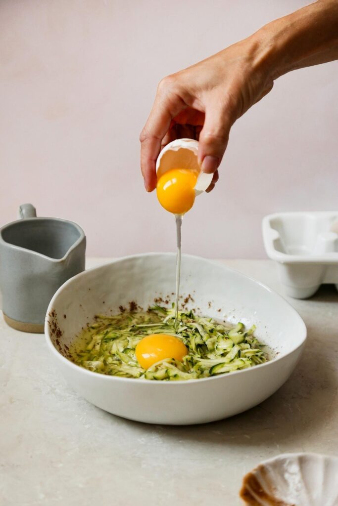 Side view of zucchini oatmeal bread wet ingredients in a white bowl. An egg id being cracked into the bowl.