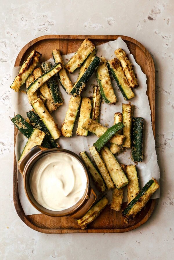 Overhead view of air fryer zucchini fries on wooden board with garlic aoili.