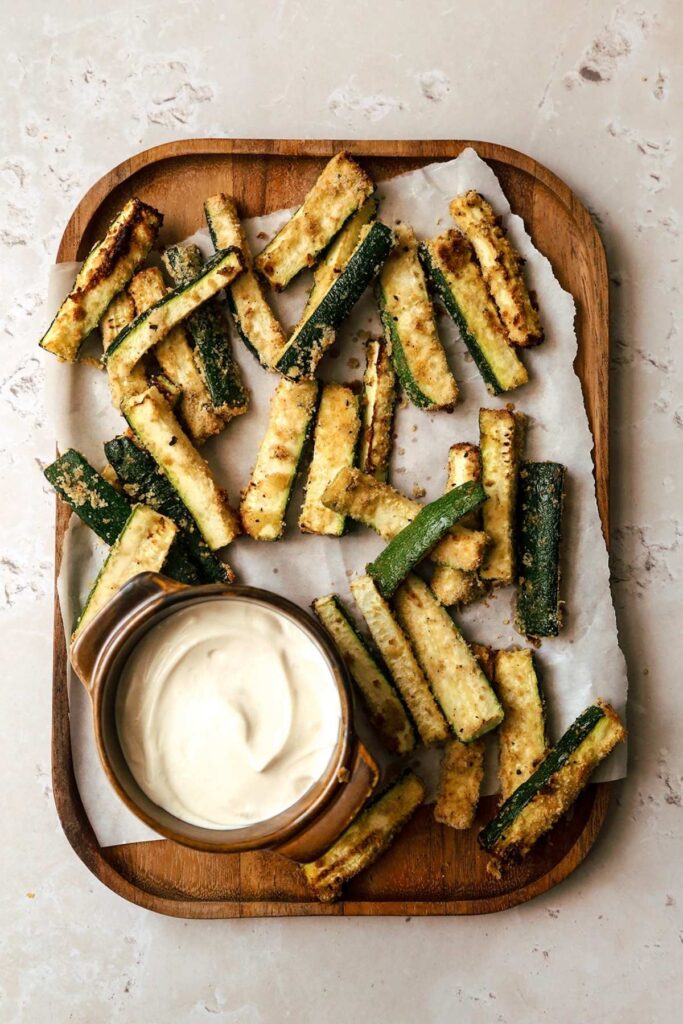 Overhead view of air fryer zucchini fries on wooden board with garlic aoili.