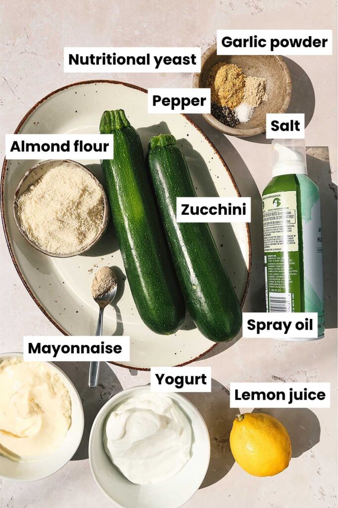 Overhead ingredient image that includes the following ingredients: zucchini, nutritional yeast, garlic powder, pepper, salt, almond flour, spray oil, mayonnaise, yogurt, and lemon juice.