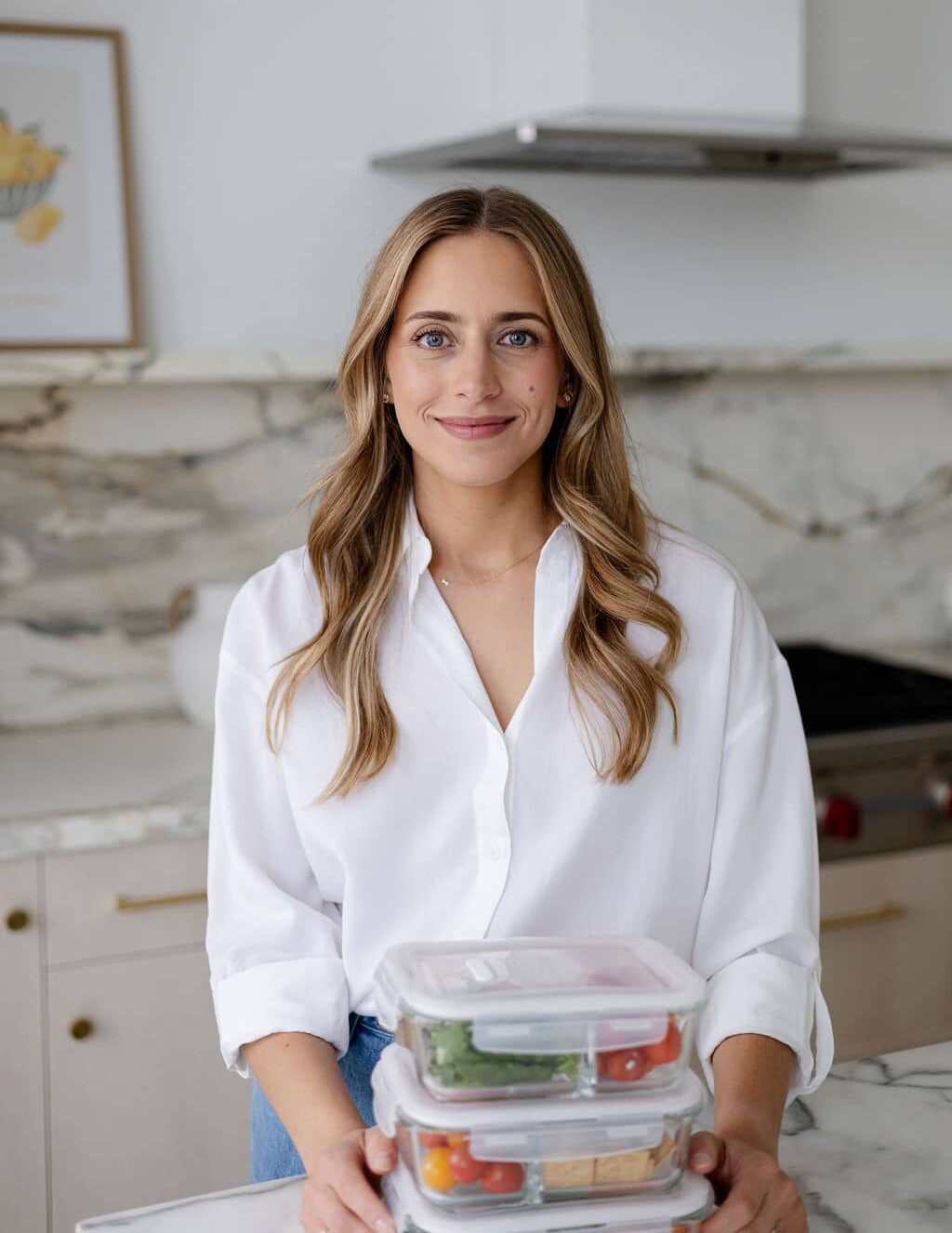 Woman in white button up standing at a counter with glass containers of meal prep food in kitchen.