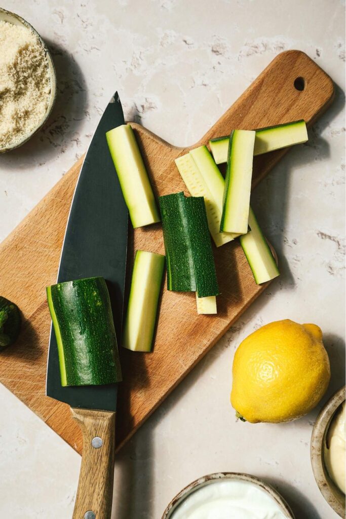 Cut zucchini on a wooden cutting board with ingredients on the side.